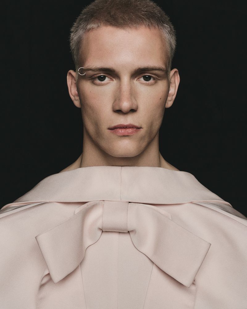 Portrait of a light-skinned, short-haired male model with an eyebrow ring, wearing a pale pink top with a large bow against a dark background.