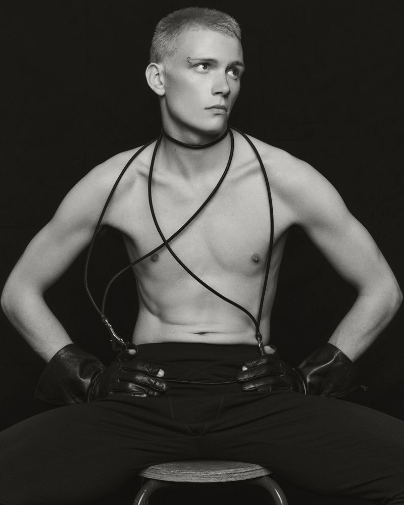 Monochrome portrait of a bare-chested adult male sitting on a stool, wearing gloves and a cord around his neck, against a dark backdrop.