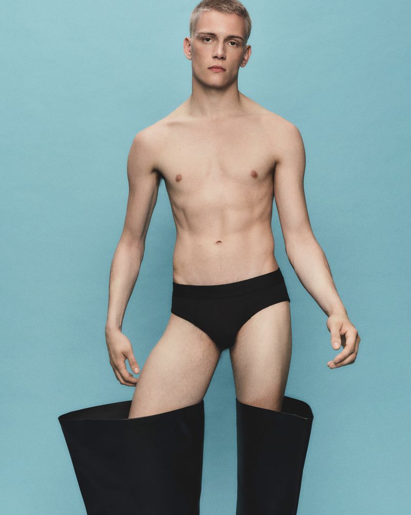 Young man poses shirtless in underwear and black rubber boots against a blue backdrop in a fashion-style photograph, eye-level shot.