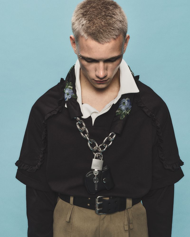 A young adult male poses indoors, wearing a black ruffled-trim top, belted tan pants, and silver chain with a padlock against a blue background.