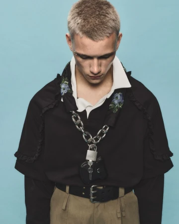A young adult male poses indoors, wearing a black ruffled-trim top, belted tan pants, and silver chain with a padlock against a blue background.