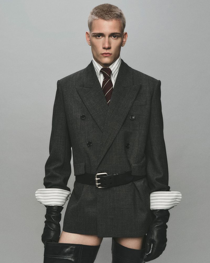 A fair-skinned man with a buzzed blonde haircut posing in a grey suit jacket, striped shirt, burgundy tie, black leather belt, gloves, and boots.