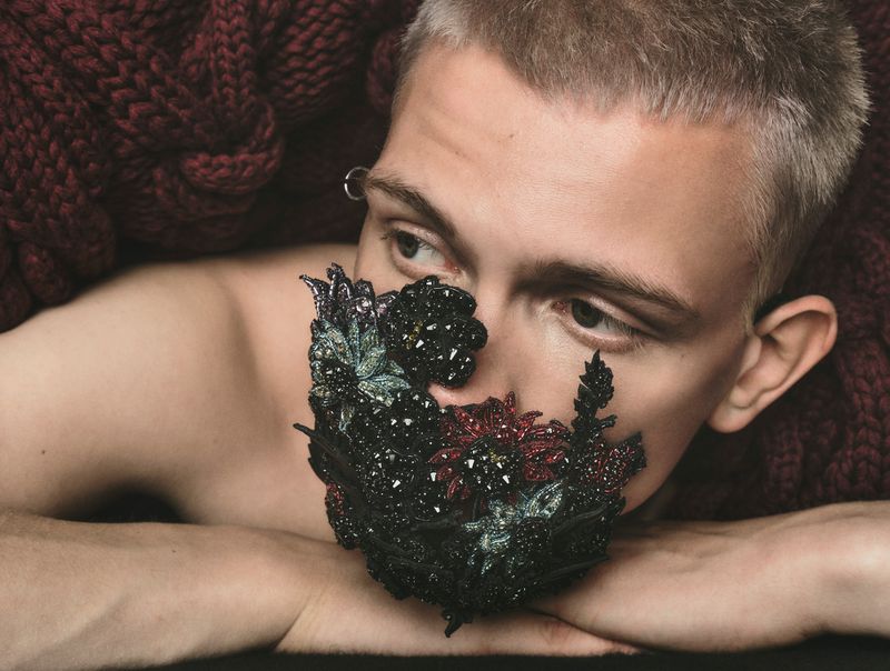 Close-up portrait of a person with short hair wearing a decorative mask, posing with their arms crossed on a dark red knitted blanket.