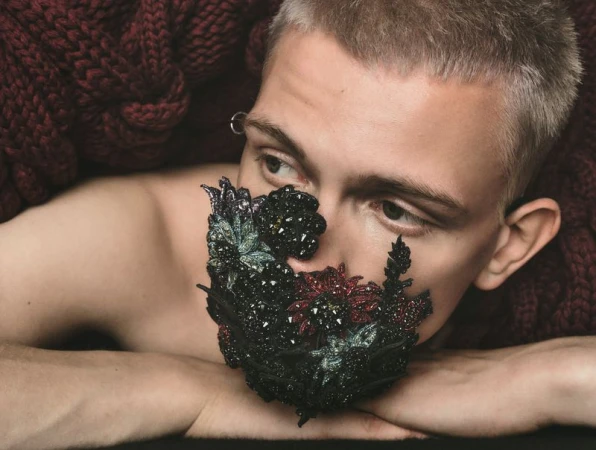 Close-up portrait of a person with short hair wearing a decorative mask, posing with their arms crossed on a dark red knitted blanket.