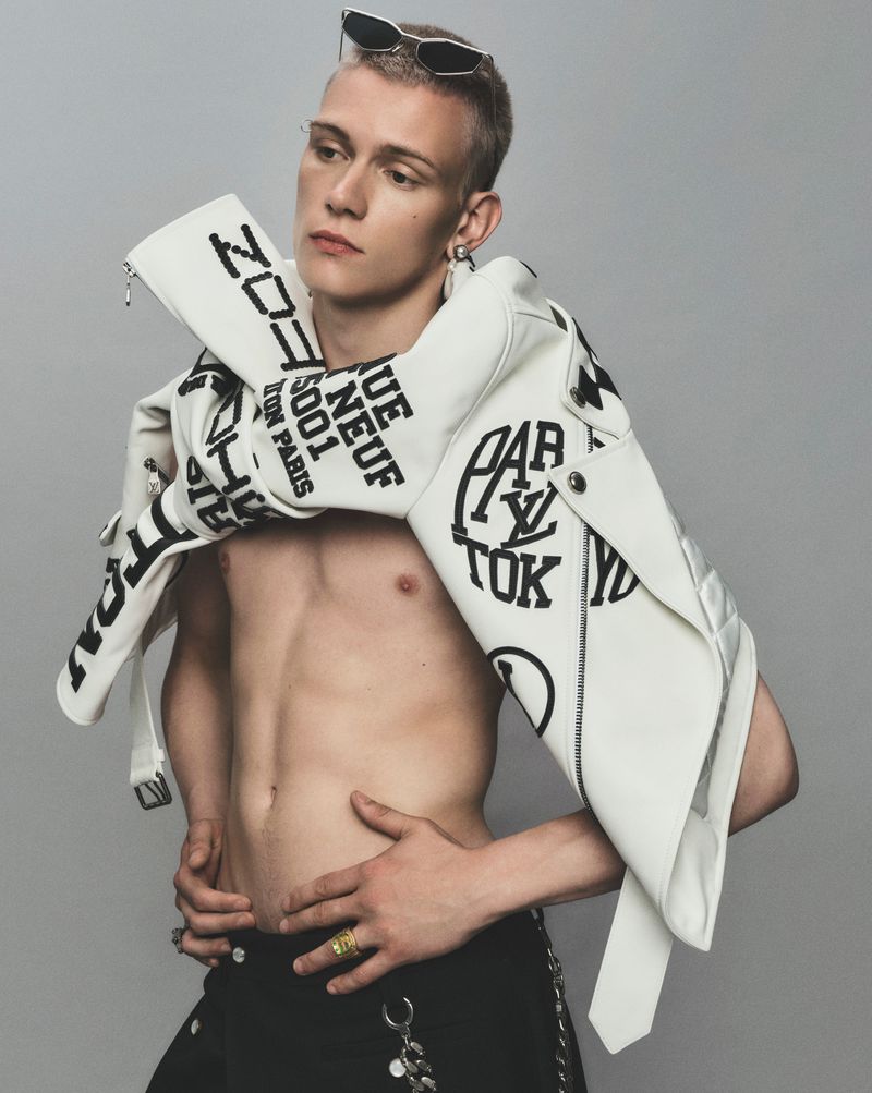 Fashion shot of a young adult male model with sunglasses, wearing a jacket with bold typography, against a neutral grey studio backdrop.