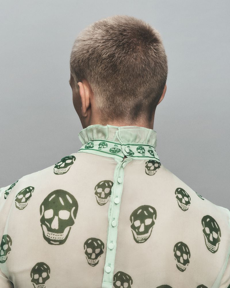Back view of a person with a short blonde haircut wearing a semi-sheer blouse adorned with a skull pattern against a gray background.