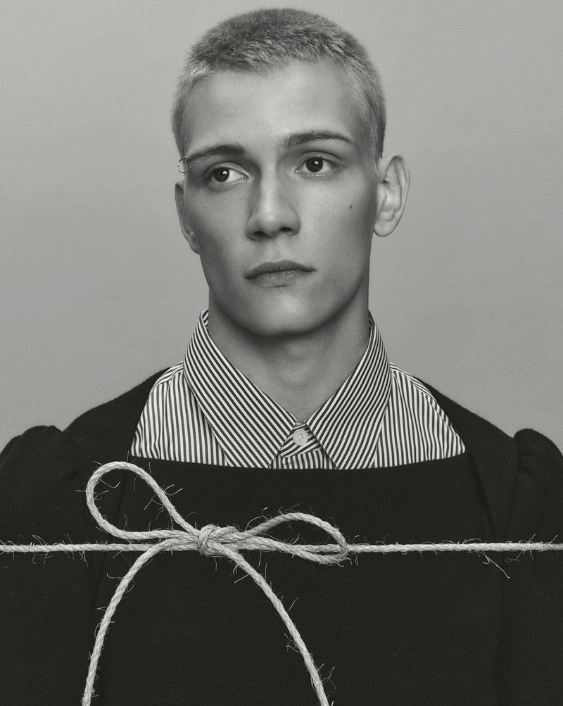 Monochrome portrait of a young adult male with short blond hair wearing a striped shirt and dark sweater with string tied across his chest.