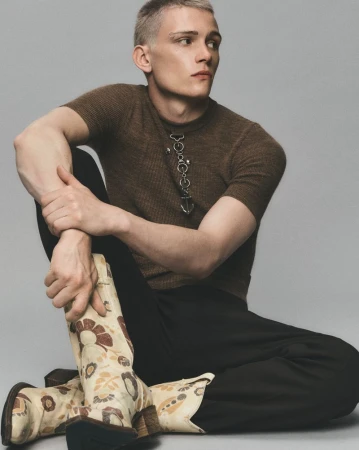 Man in brown shirt and black pants sits on the floor with floral cowboy boots in a studio environment.