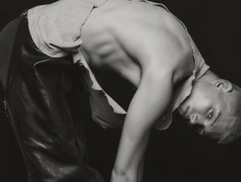 Monochrome portrait of model Will Woodward arching backward wearing a torn sleeveless top and leather pants against a black background.