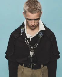 Close-up portrait of model Will Woodward wearing a black top with floral embroidery, chunky chain necklace, and padlock accessory against blue.