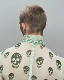Rear view of a person wearing a sheer top with green skull print and ruffled collar against a grey background.