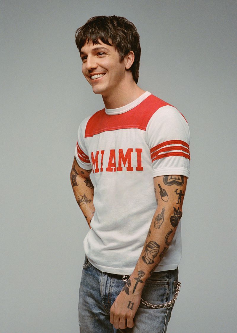 Smiling adult male in studio shot wearing a red and white shirt with 'Miami' printed on it, blue jeans, and has visible tattoos on arms.