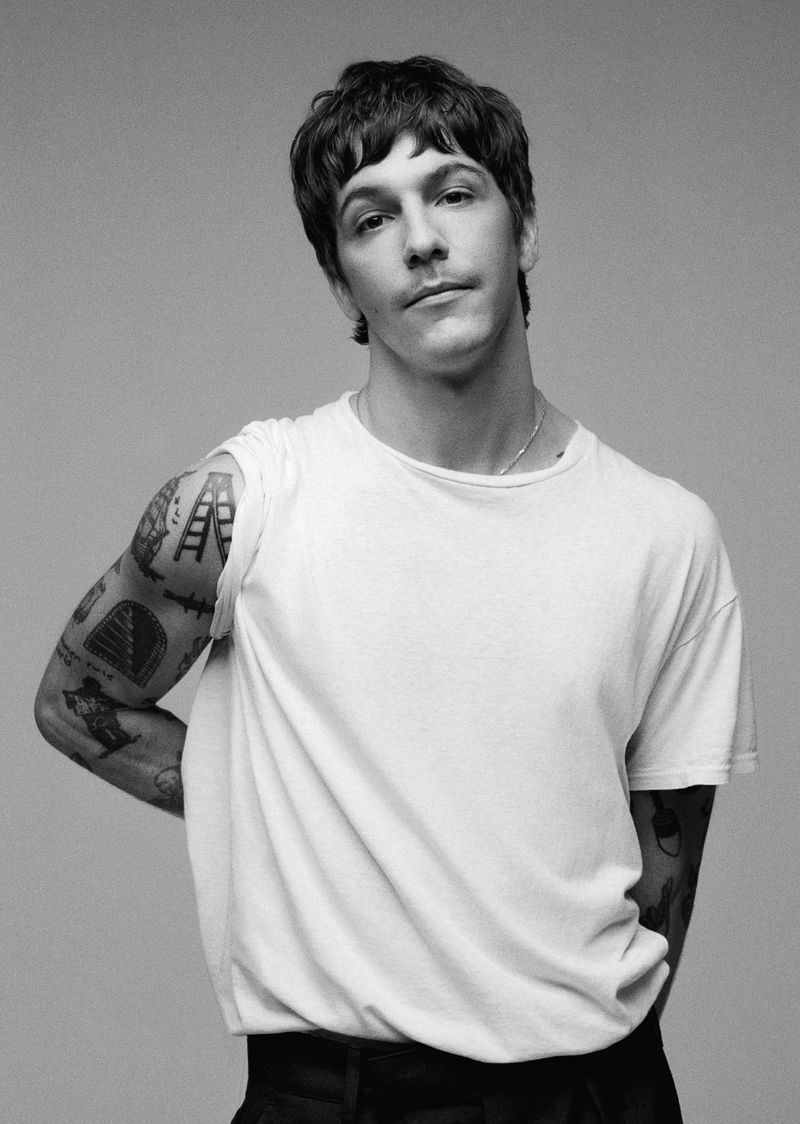 Black-and-white portrait of an adult male with dark hair in a white t-shirt, posing in a studio with tattoos on his left arm.