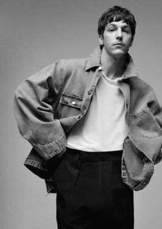 Portrait of a young adult in a denim jacket, white t-shirt, and black pants, posing in a studio with a neutral gray background.
