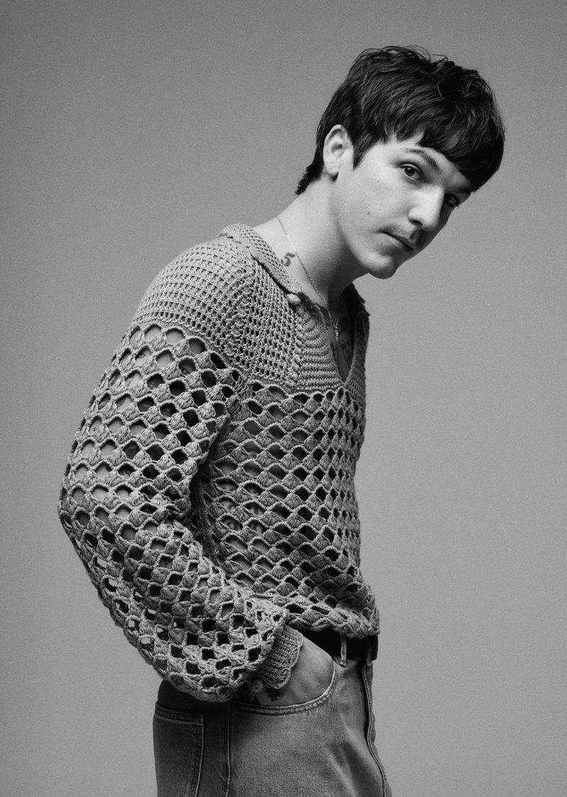 Studio shot of a young adult man wearing a crochet sweater and jeans, posing against a plain background in black and white.