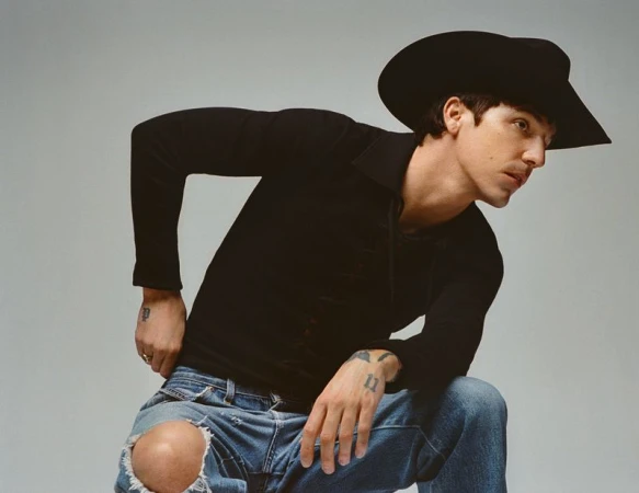 Portrait of a man in a black long-sleeve shirt, cowboy hat, and distressed jeans sitting against a neutral studio backdrop.