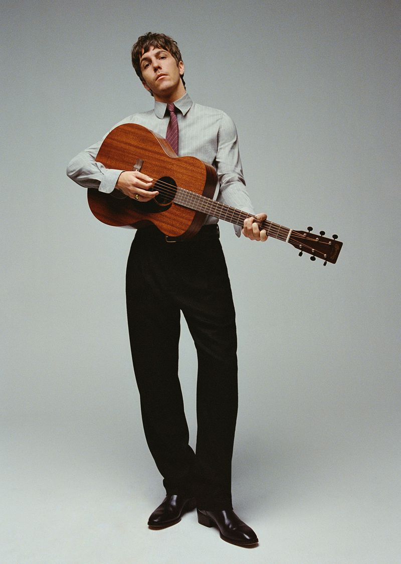 Adult male playing acoustic guitar in studio setting with professional lighting and wearing button-down shirt and dark tie.
