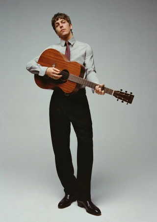 Adult male playing acoustic guitar in studio setting with professional lighting and wearing button-down shirt and dark tie.