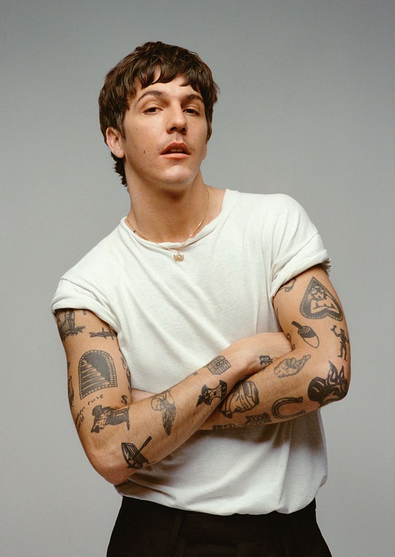 Studio portrait of a young adult male with tattoos on his arms, wearing a white t-shirt and black pants, with a neutral facial expression.