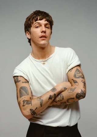 Studio portrait of a young adult male with tattoos on his arms, wearing a white t-shirt and black pants, with a neutral facial expression.