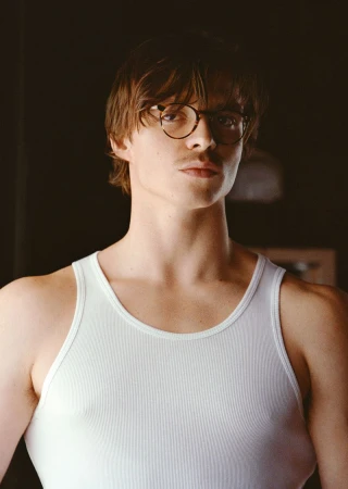 Portrait of an adult male with fair skin, wearing a white tank top and glasses, with a neutral expression in a softly lit indoor setting.