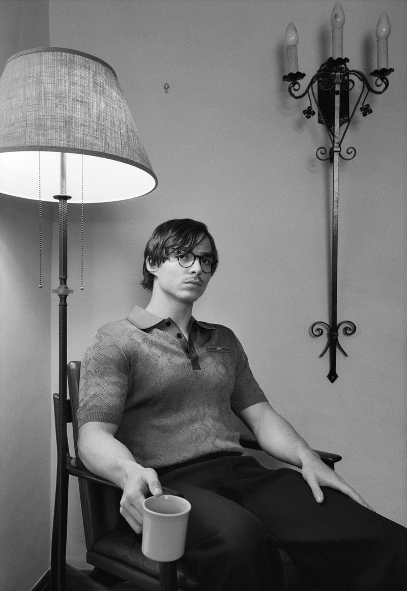 Monochrome portrait of a man with glasses sitting in a chair, holding a cup, illuminated by a floor lamp indoors.