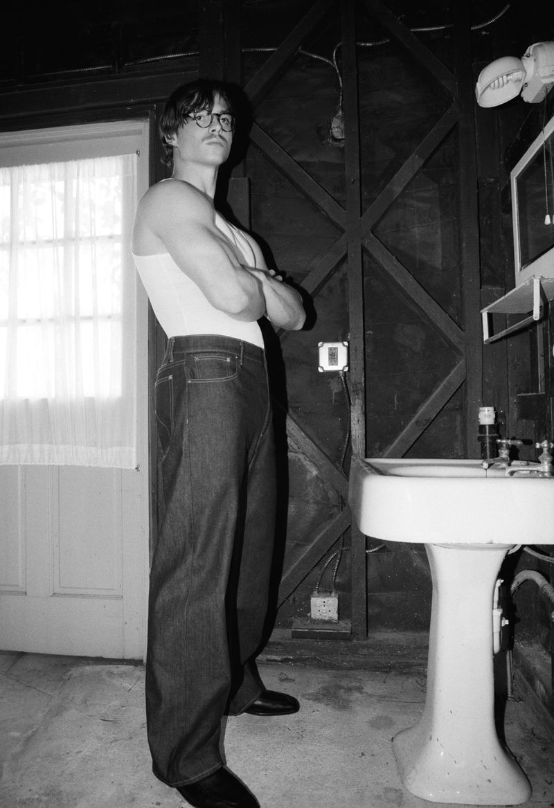 Monochrome portrait of a man in a white tank top and jeans, standing indoors with arms crossed, captured with a shallow depth of field.