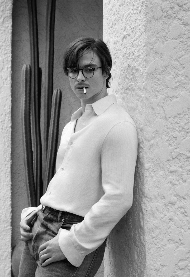 Black-and-white portrait of an adult man wearing glasses and holding a cigarette in his mouth, posing outdoors with a cactus plant.
