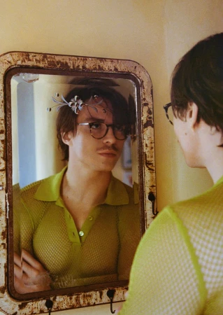 Man in green shirt looking in a vintage mirror indoors with hooks and cream wall in the background, vintage effect filter applied.