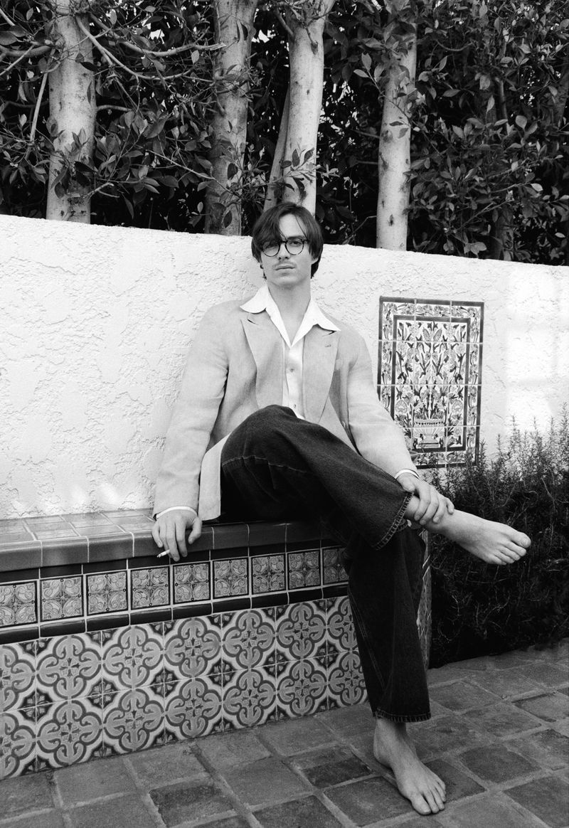 Black and white portrait of a man sitting outdoors on a tiled bench. He is wearing glasses, a blazer, jeans, and is barefoot.