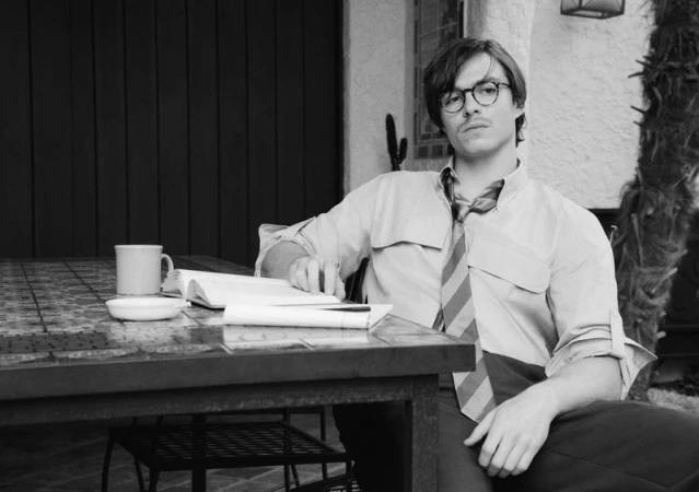 Monochrome portrait of a man with glasses seated at a table, reading a book, outdoors with a tiled surface and neutral wall behind.
