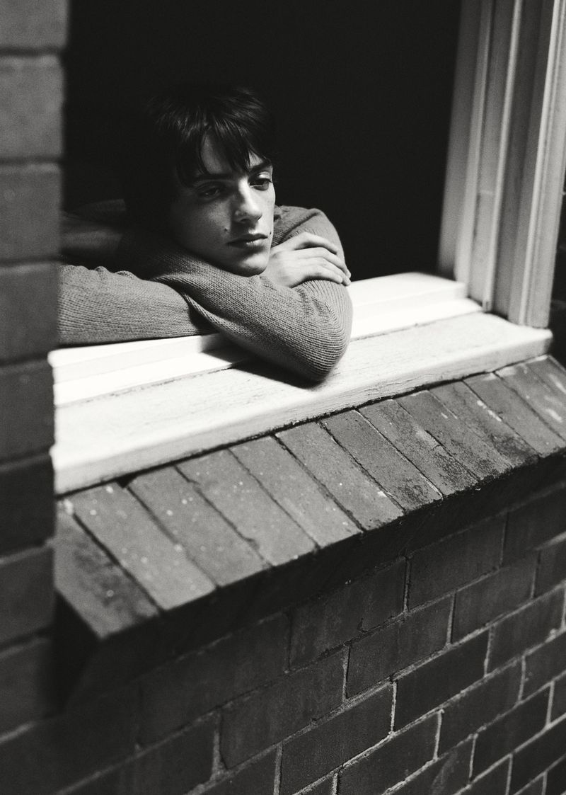 Portrait of a contemplative young adult man in a sweater, resting his arms on a windowsill, captured in a black and white photo.