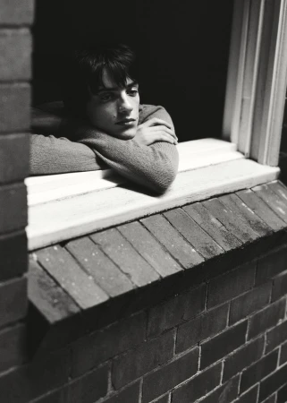 Portrait of a contemplative young adult man in a sweater, resting his arms on a windowsill, captured in a black and white photo.