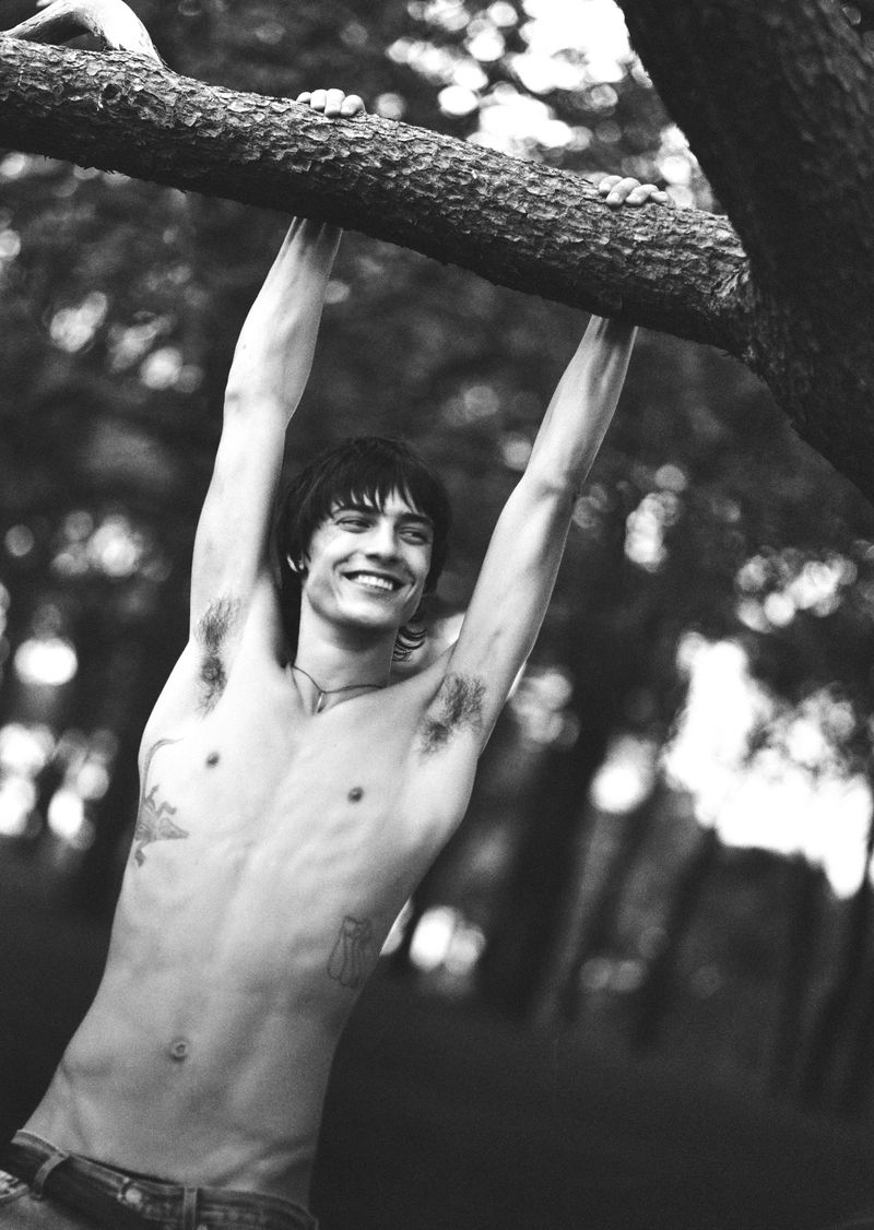Black and white portrait of a shirtless young adult male outdoors, hanging from a tree branch with a smile, set against a natural background.