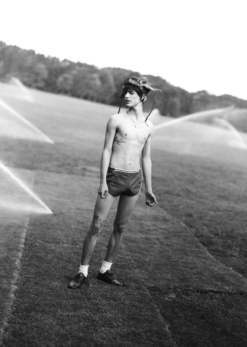 Black and white photo of a shirtless young man in athletic shorts standing on a grassy hill with sprinklers in the background.