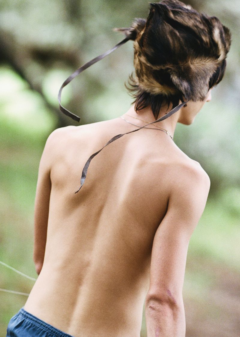 Back view of a person with bare torso wearing a feathered headdress and blue shorts outdoors in natural light and shallow depth of field.