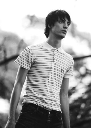 Black and white portrait of a young adult male wearing a striped polo shirt, standing outdoors against a blurred background.