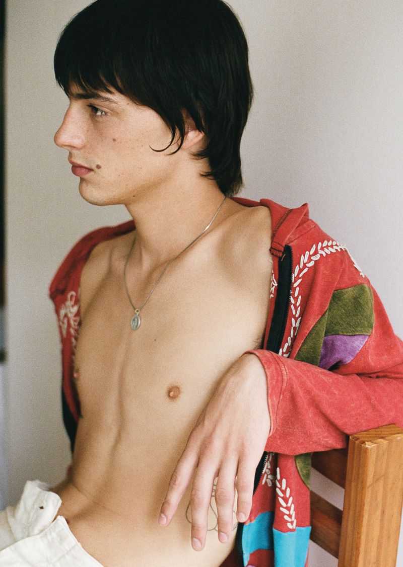 Side profile portrait of a young adult male with dark hair, wearing an open, patterned shirt, showcasing a natural and relaxed aesthetic.