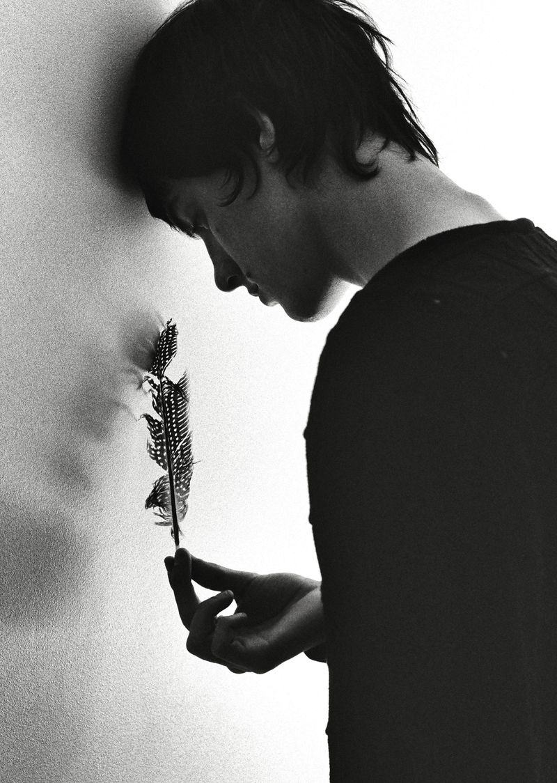 Monochrome portrait of a young adult leaning against a wall and pensively holding a spotted feather in hand, indoor setting.