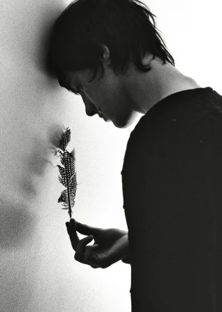 Monochrome portrait of a young adult leaning against a wall and pensively holding a spotted feather in hand, indoor setting.
