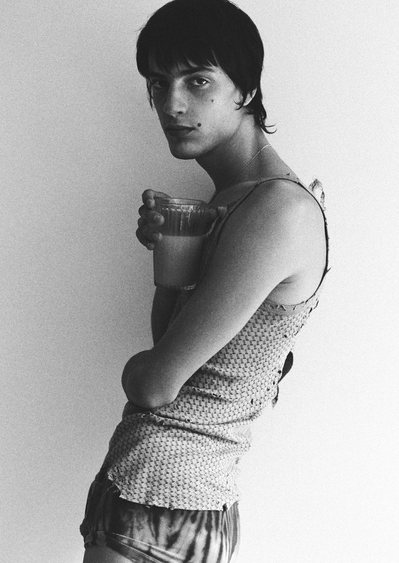 Monochrome portrait of a slender adult with dark hair holding a clear glass of a milky-beige liquid in a simple indoor setting.