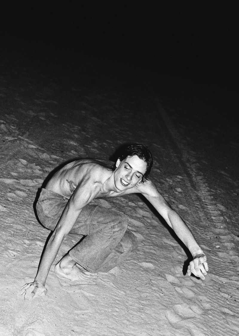 Monochrome portrait of a shirtless young adult on a sandy slope at night, with arms outstretched and a smiling expression.