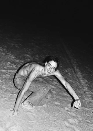 Monochrome portrait of a shirtless young adult on a sandy slope at night, with arms outstretched and a smiling expression.