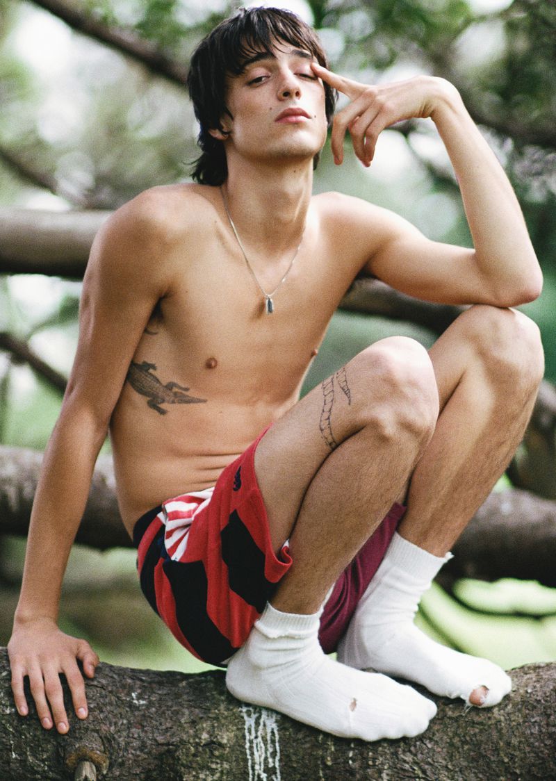 A shirtless young man with dark hair and a tattoo squats on a tree branch wearing socks and red and black shorts in a natural setting.