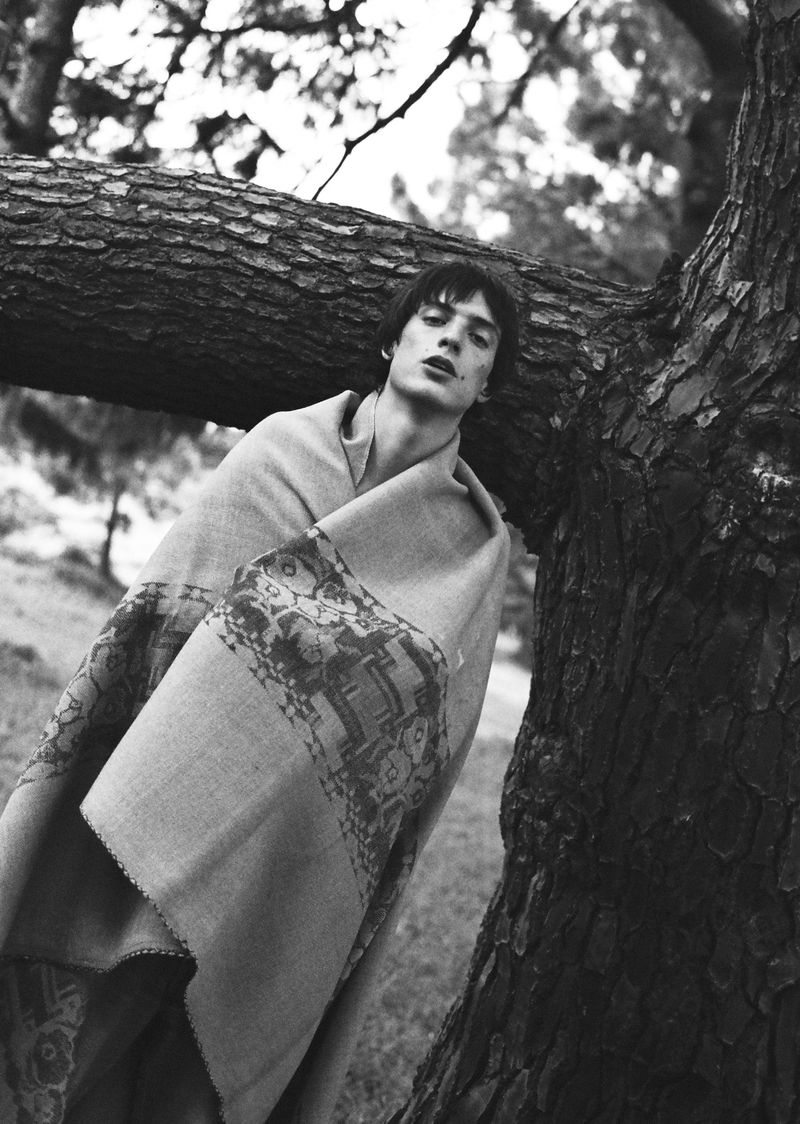 Black and white portrait of a young adult male wrapped in patterned blanket, leaning against a tree, set in a natural outdoor environment.