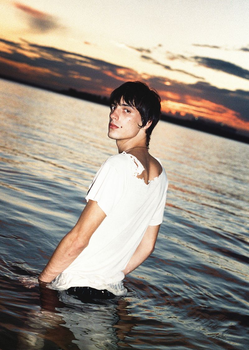 Young adult stands waist-deep in water against a vibrant sunset sky with orange hues reflecting in the tranquil water surface.