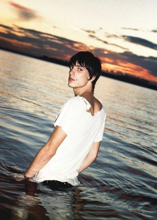 Young adult stands waist-deep in water against a vibrant sunset sky with orange hues reflecting in the tranquil water surface.