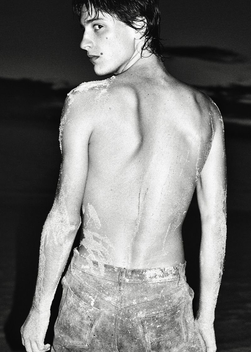 Black and white shot of a shirtless young man with sandy skin and wet hair looking over his shoulder against a dark outdoor backdrop.