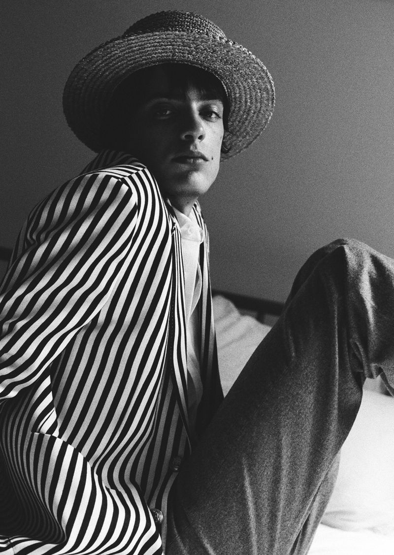 Monochrome portrait of a stylish adult male with straw hat, striped jacket, and jeans, posing indoors with a contemplative gaze.