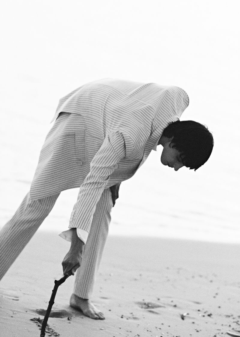 Black and white photo of a slim adult male in a suit bending over on a sandy beach, holding a stick and drawing in the sand.